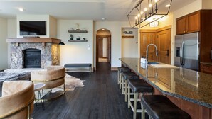 A modern kitchen with a stone fireplace, bar stools, and a large island with a stainless steel refrigerator.