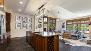 A spacious kitchen with a large island, wooden cabinets, and a wine rack. There is a dining area with a gray sofa and a rug.