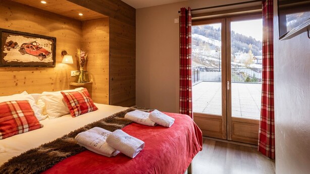 A hotel room with a wooden interior, a bed with red bedding, a window with a view of a snowy landscape, and a painting on the wall. - 4-room apartment, 8 people, pets allowed, terrace (Valloire)