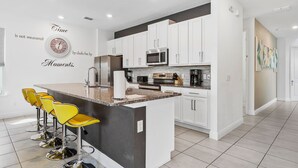 A modern kitchen with white cabinets, a central island with bar stools, and a wall clock. - Liberty Hope: Private Pool, Water Park, Golf View, Near Disney (Kissimmee)