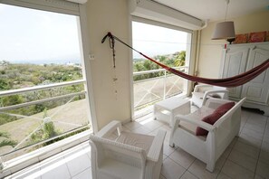 A balcony with white wicker furniture, a hammock, and a view of greenery and buildings. - 5 minutes from Anse Mitan beach, quiet apartment with sea view (Les Trois-Îlets)