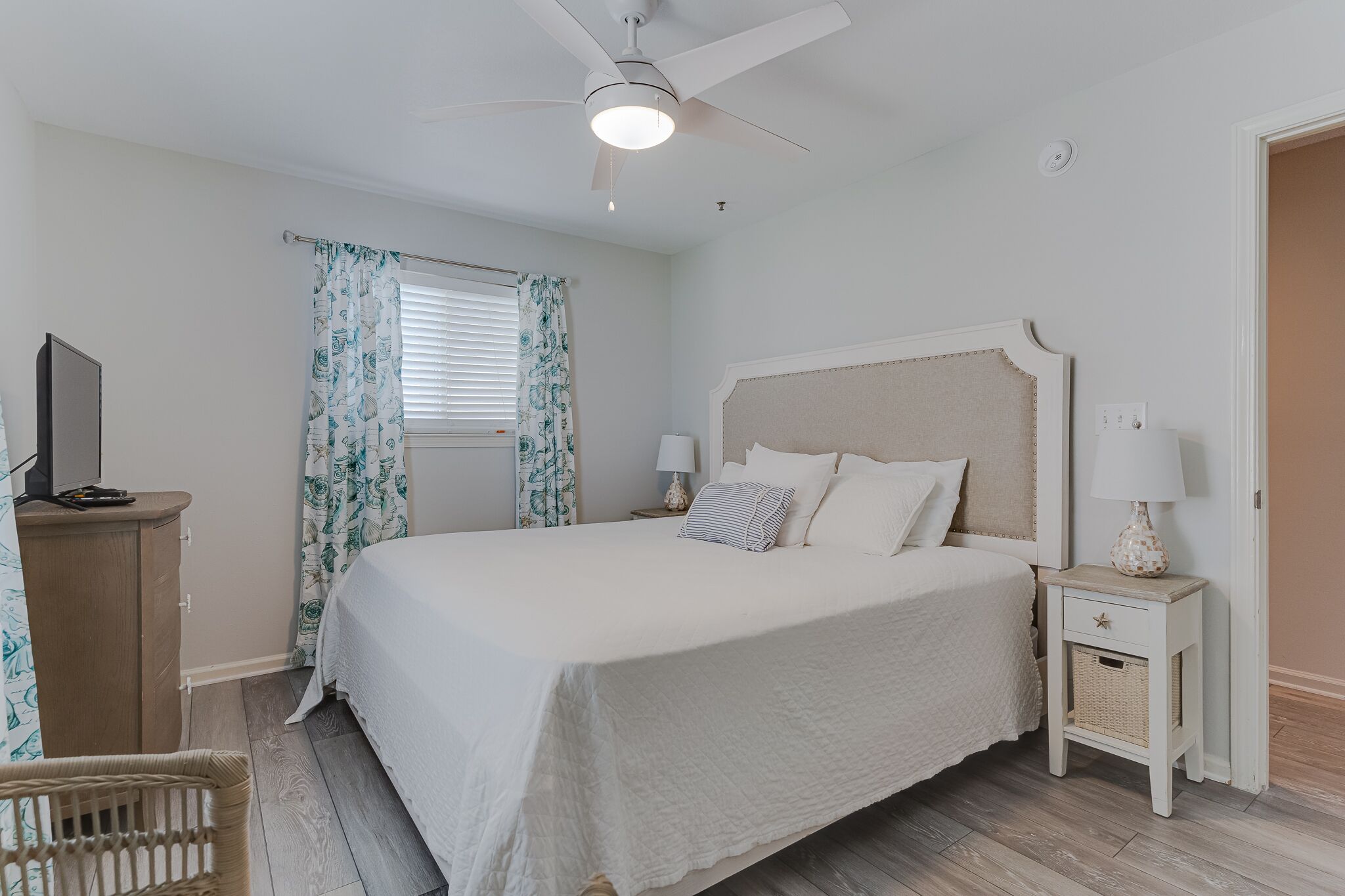 Une chambre avec un grand lit, un ventilateur de plafond, une table de chevet avec une lampe, un téléviseur posé sur une commode en bois et un panier en osier.