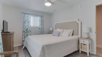 Une chambre avec un grand lit, un ventilateur de plafond, une table de chevet avec une lampe, un téléviseur posé sur une commode en bois et un panier en osier.
