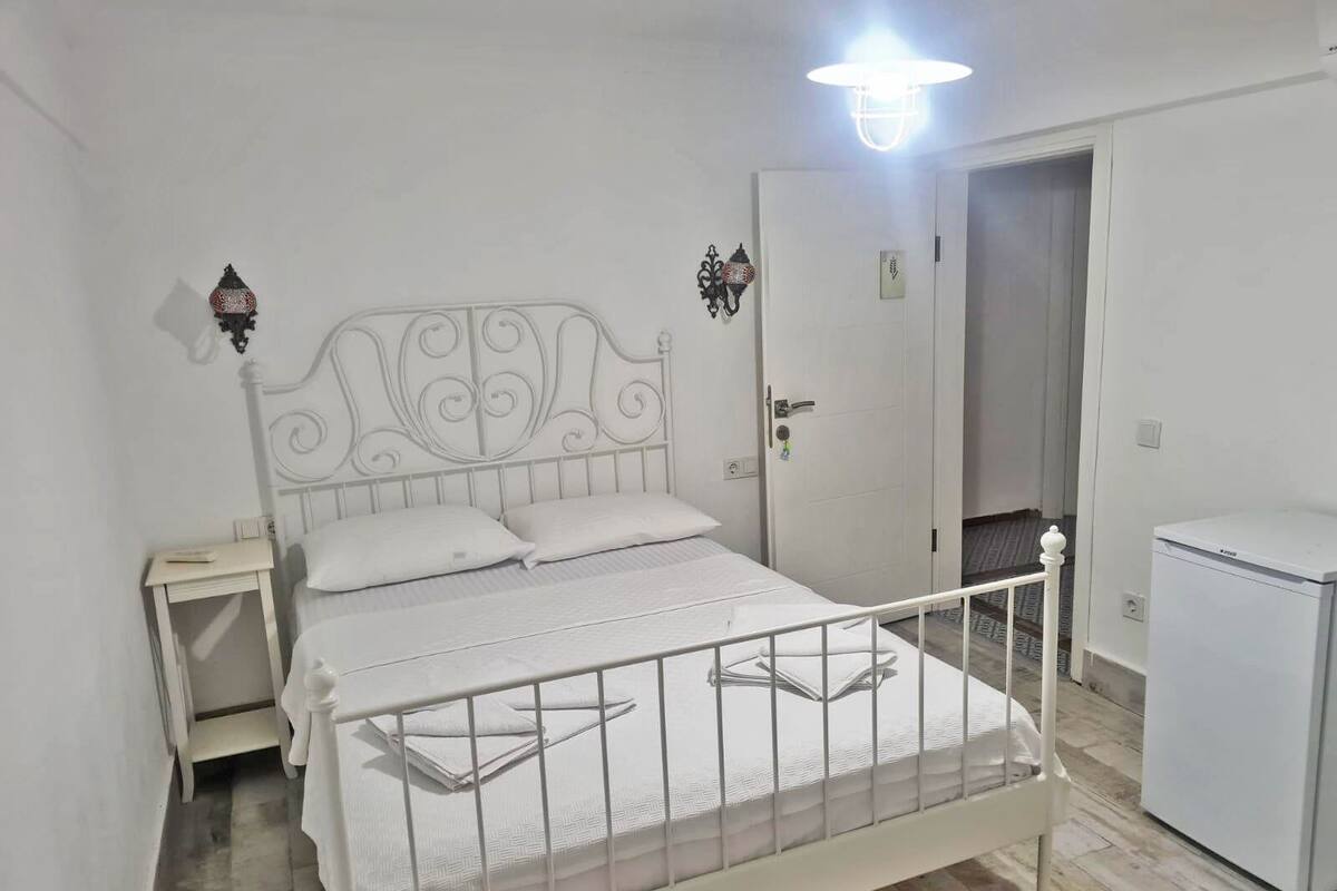 Una cama de metal blanco con cabecera decorativa, una mesita de noche, una lámpara fijada a la pared y un refrigerador blanco.