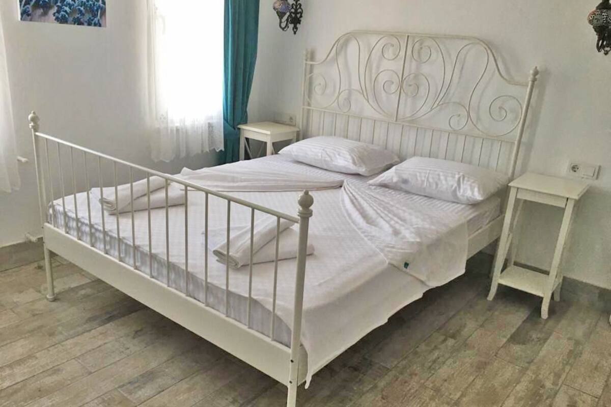 Una cama de metal blanca con cabecera, dos mesitas de noche y una ventana con cortinas.