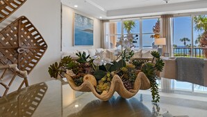 Un salon moderne avec une table basse en verre, des fauteuils en osier, un ventilateur de plafond et une vue sur l’océan grâce à de grandes fenêtres.