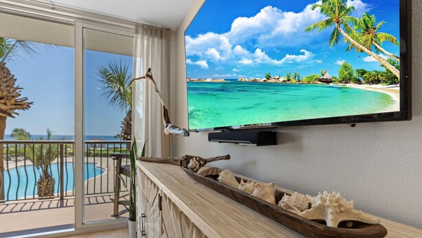 Un balcon avec une piscine, une petite table d’appoint en bois ornée de coquillages et un téléviseur diffusant une scène de plage.