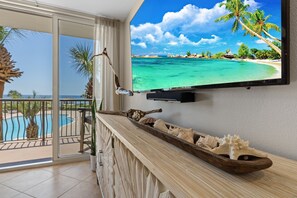 A balcony with a pool, a wooden side table with seashells, and a TV displaying a beach scene. - Stunning Remodel, Gorgeous Gulf Views, Private Beach - BEACH SERVICE INCLUDED (Miramar Beach)