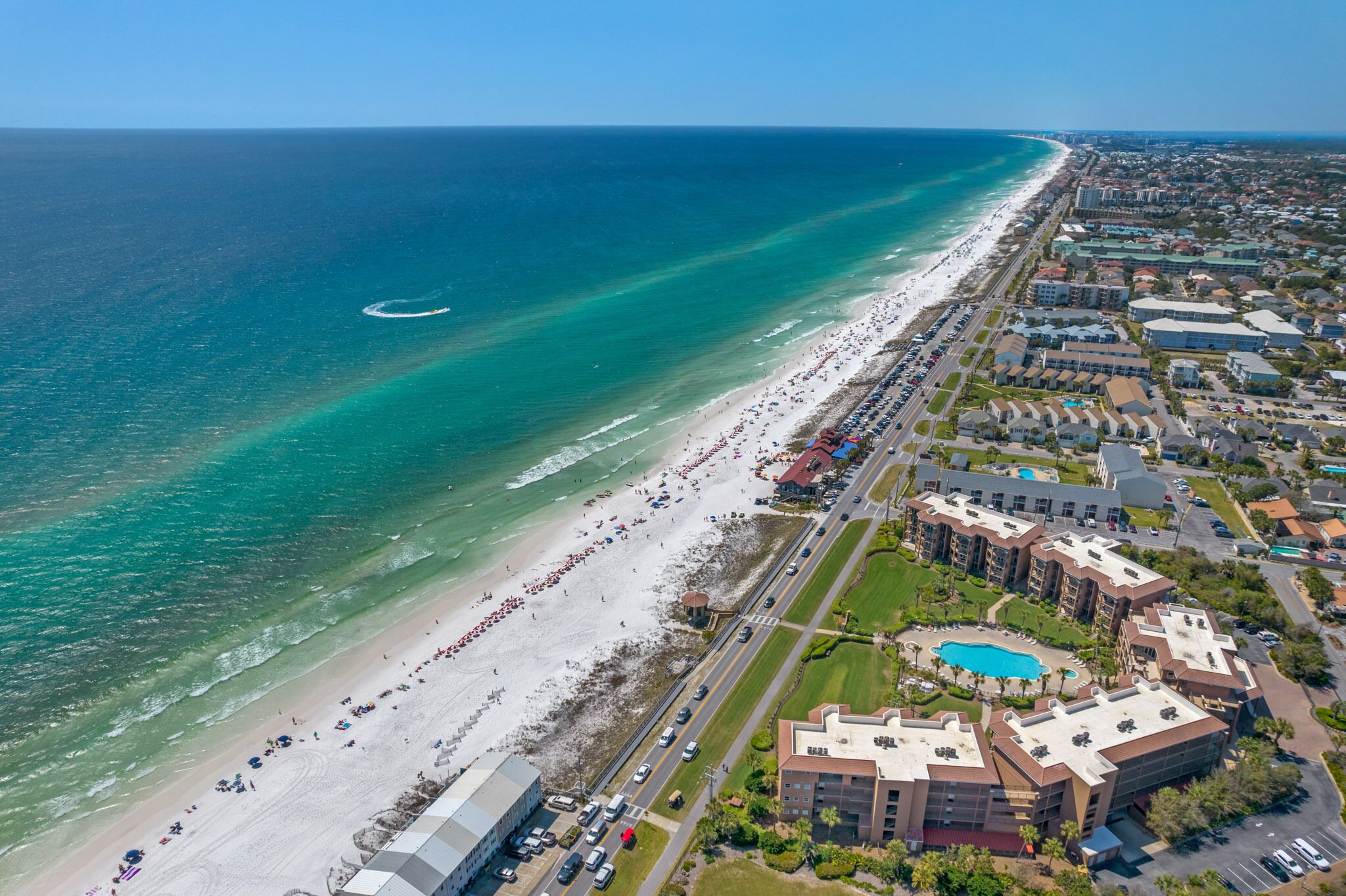 An aerial view of a coastal area with a beach, a swimming pool, and adjacent residential buildings.