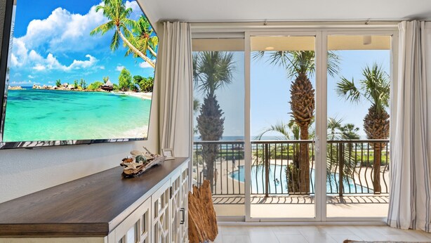A room with a TV displaying a beach scene, a wooden counter, a glass door leading to a balcony with a pool and palm trees. - The ONLY 1 Bedroom with a private Bunk Room - Beach Service Included!!! (Miramar Beach)
