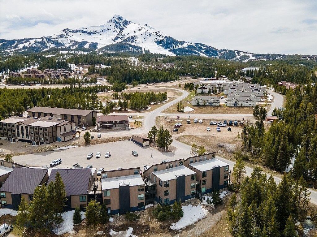 A multi-story building with a parking lot in front, surrounded by trees and a mountain in the background.