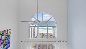 Une pièce dotée d’une grande fenêtre en arc donnant sur une plage, d’un ventilateur de plafond et d’un tableau abstrait aux couleurs vives accroché au mur.