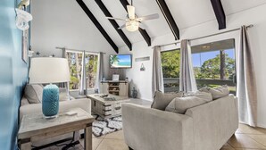 Ein geräumiges Wohnzimmer mit einer großen Couch, einem Couchtisch und einem Fernseher. Der Raum verfügt über eine hohe Decke mit freiliegenden Balken und große Fenster mit Vorhängen.