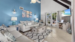Ein Wohnzimmer mit blauer Wand, einem Deckenventilator, einem Fernseher, einem weißen Sofa, einem Couchtisch und Blick ins Freie durch eine Glastür.