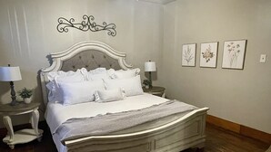 Una cama bien hecha con cabecera, dos mesitas de noche con lámparas, y tres impresiones botánicas enmarcadas en la pared.