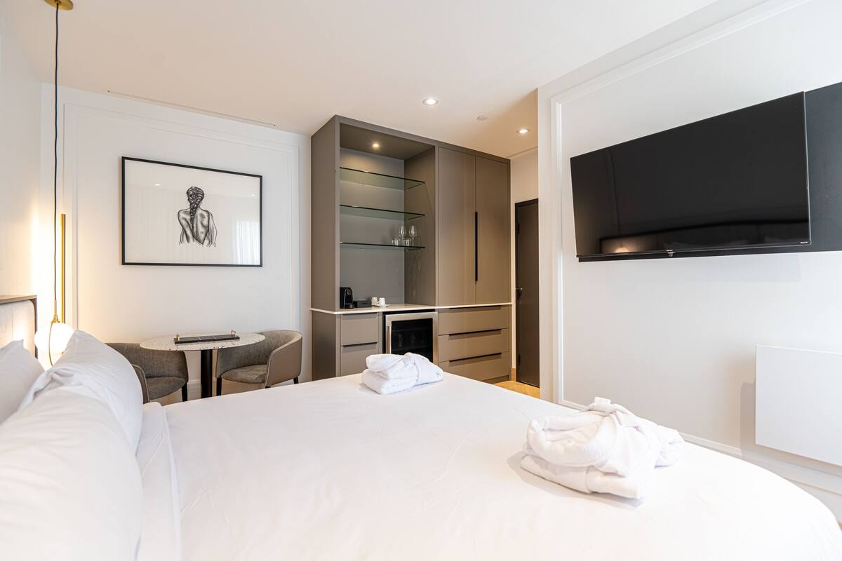 A modern hotel room with a large bed, a flat-screen TV, a small desk with a chair, and a glass shelving unit.