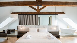 Una habitación de hotel moderna con una cama grande, techo de madera, tragaluz, bañera y una zona de estar.
