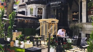 Un área de estar al aire libre con mesas y sillas, una pérgola de madera y una persona trabajando en un mostrador.