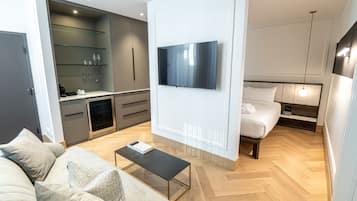 Een moderne hotelkamer met een flatscreen televisie aan de muur, een bank met kussens, een klein bijzettafeltje, een bed met hoofdbord en een ingebouwde kitchenette met een magnetron en een koffiezetapparaat.