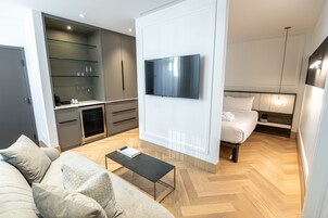 Una habitación de hotel moderna con televisión de pantalla plana en la pared, un sofá con cojines, una pequeña mesa de centro, una cama con cabecera y una cocina integrada con microondas y cafetera.