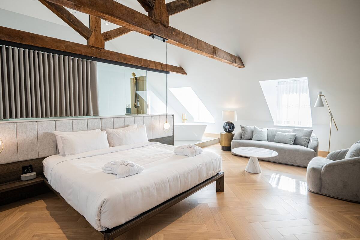 A modern hotel room with a large bed, a sofa, a small table, and a view of the city through large windows.
