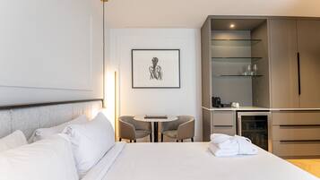 Une chambre d’hôtel comprenant un lit, une petite table ronde avec deux chaises, une œuvre d’art encadrée au mur, un minibar et une armoire.