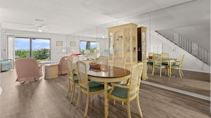 A spacious room with a dining area featuring a round table and wicker chairs, a large window with a view, and a staircase leading to an upper level. - Green Palms Oceanfront 2 Bed Townhouse (Ponce Inlet)