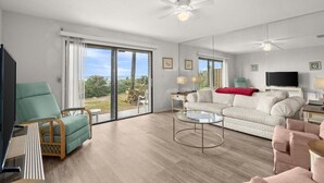 A spacious living room with a large sliding glass door leading to a balcony, a ceiling fan, a television, and a comfortable seating area. - Green Palms Oceanfront 2 Bed Townhouse (Ponce Inlet)