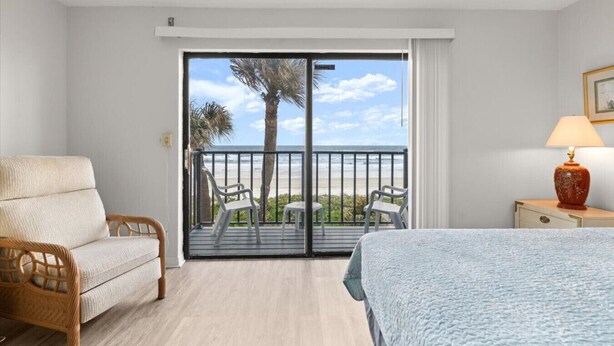 A hotel room with a balcony, a bed, a chair, a nightstand, and a lamp. - Green Palms Oceanfront 2 Bed Townhouse (Ponce Inlet)