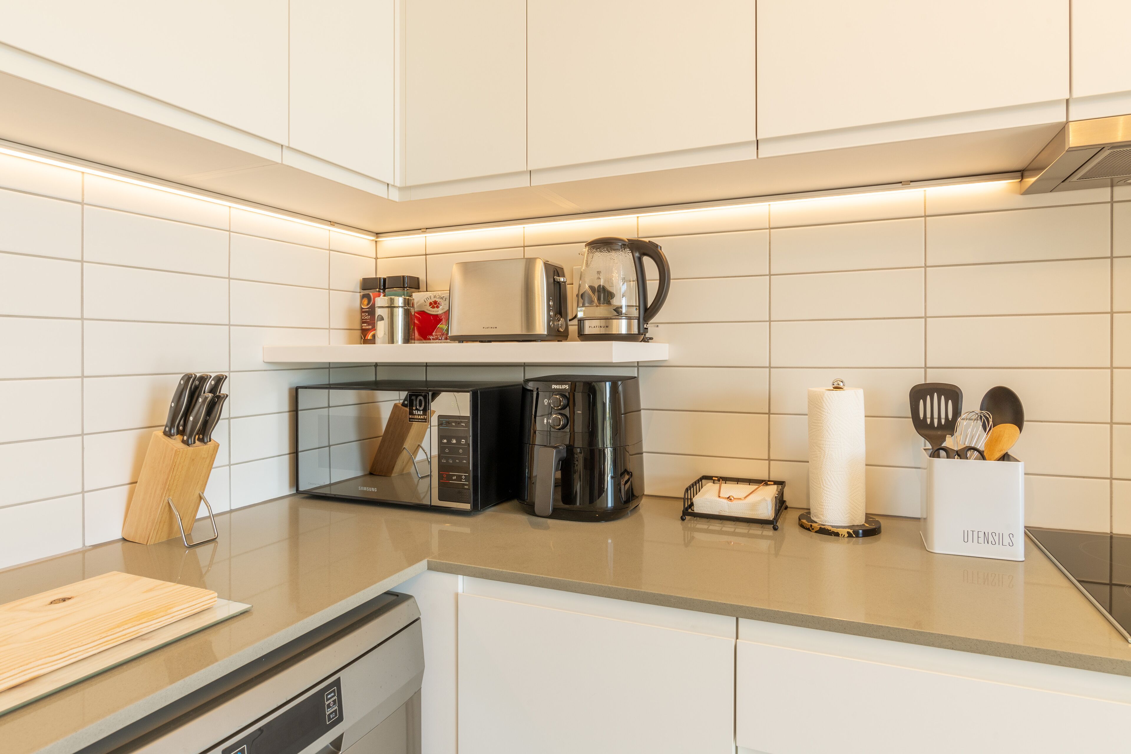 A modern kitchen with white cabinets, a microwave, a coffee maker, and a toaster. There is a knife block and a paper towel holder on the counter.