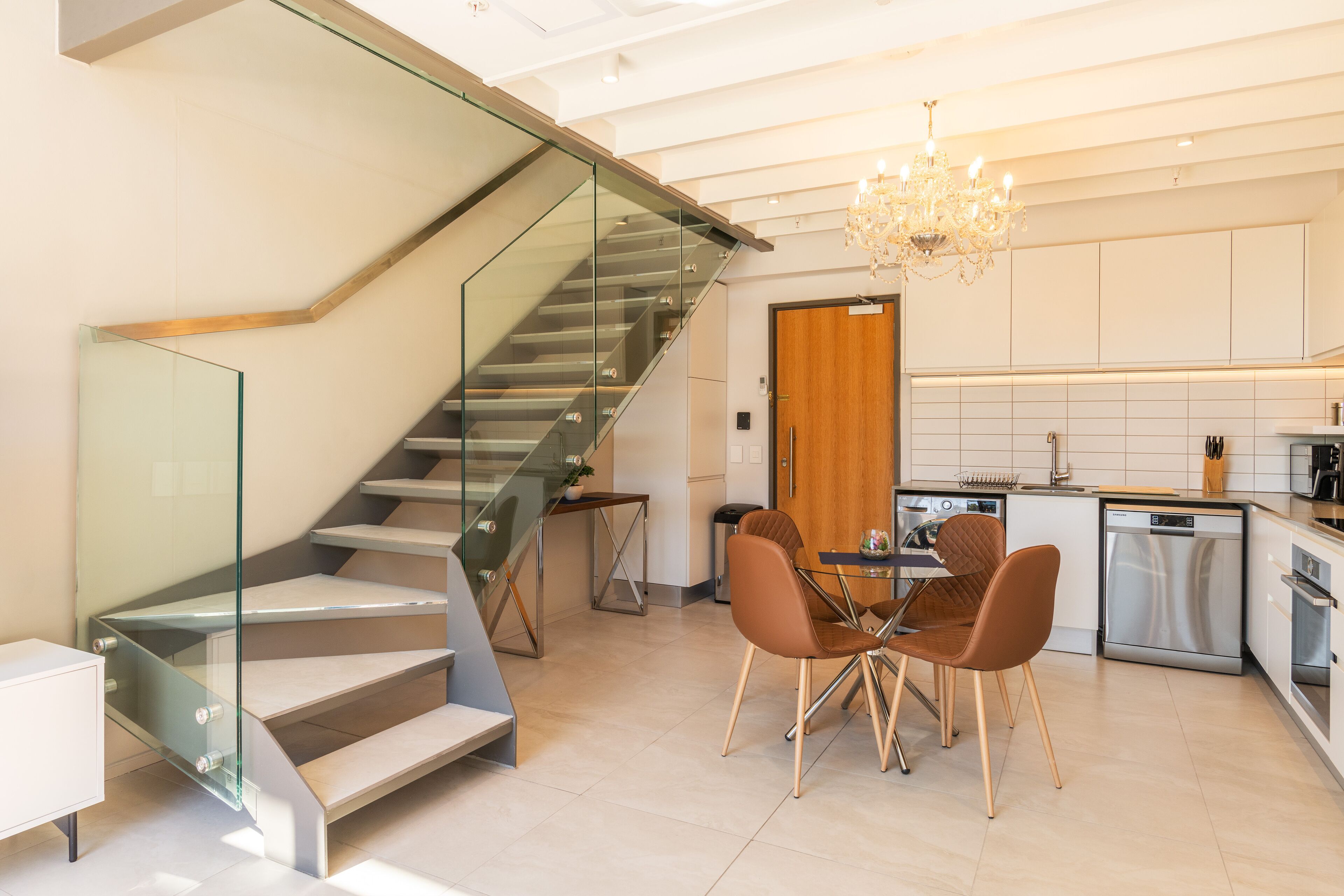 A modern kitchen with a dining area, a glass staircase, and a chandelier.