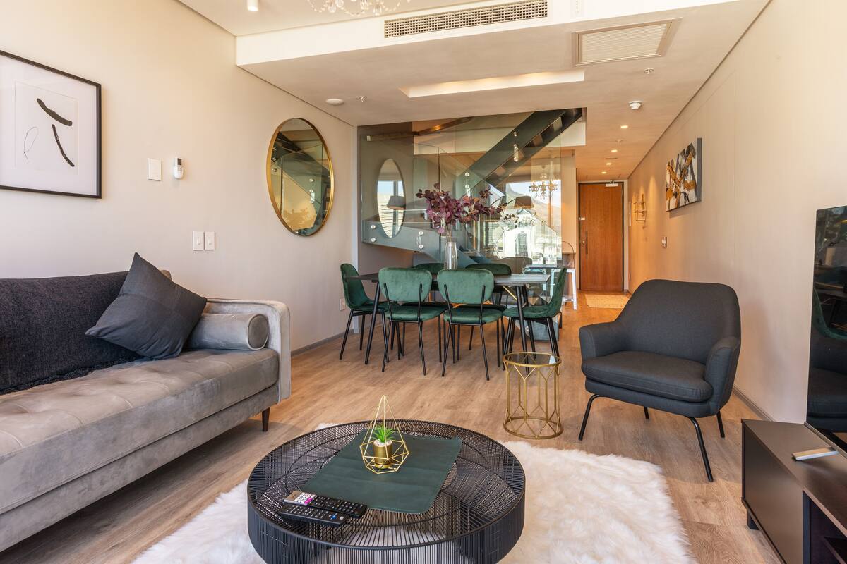 A modern living room with a grey sofa, green chairs, a round coffee table, and a staircase leading to an upper level.