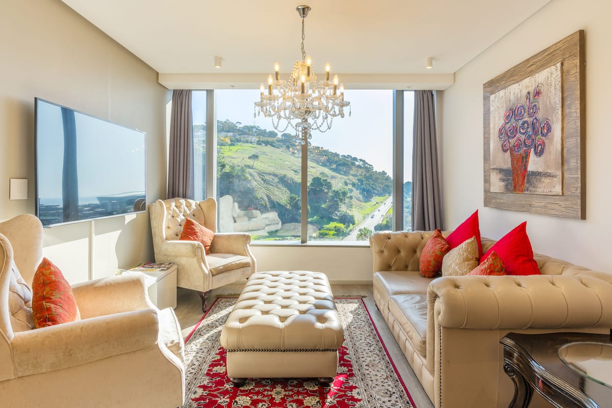 A spacious living room with a large window, a chandelier, and a painting.