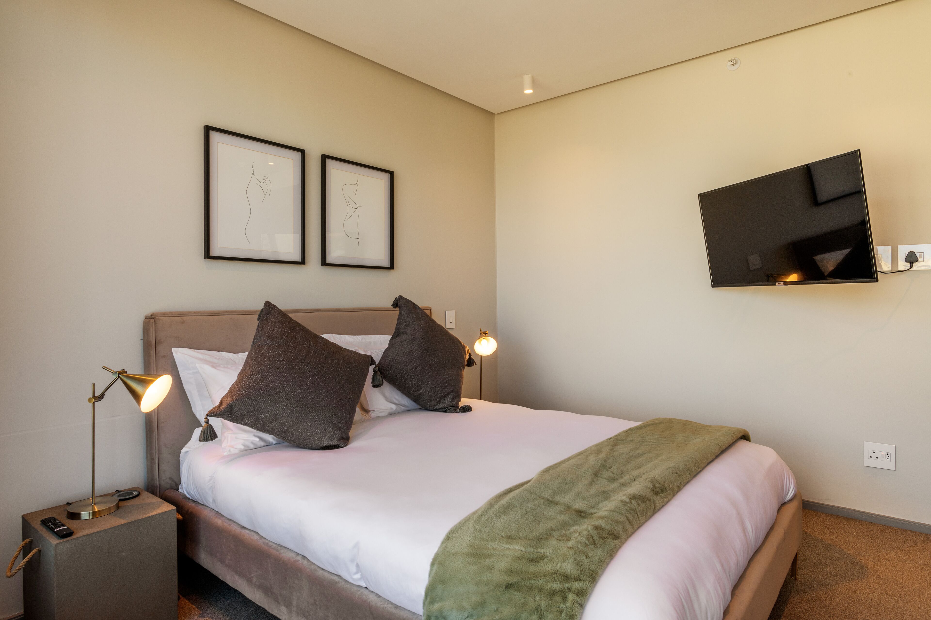A neatly made bed with a green throw blanket, two framed artworks on the wall, a bedside lamp, and a flat-screen TV mounted on the wall.