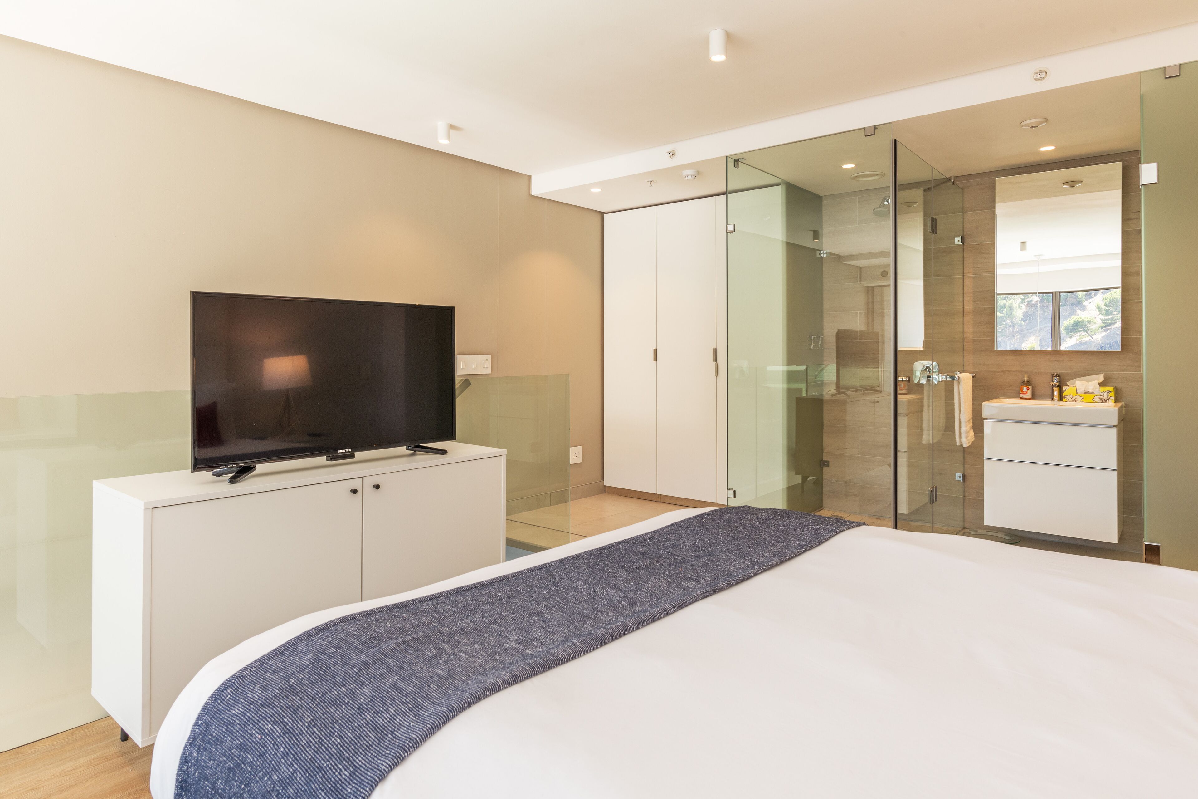 Een moderne slaapkamer met een groot bed, een televisie op een witte dressoir, een glazen douchecabine, en een badkamer met een spiegel en wastafel.