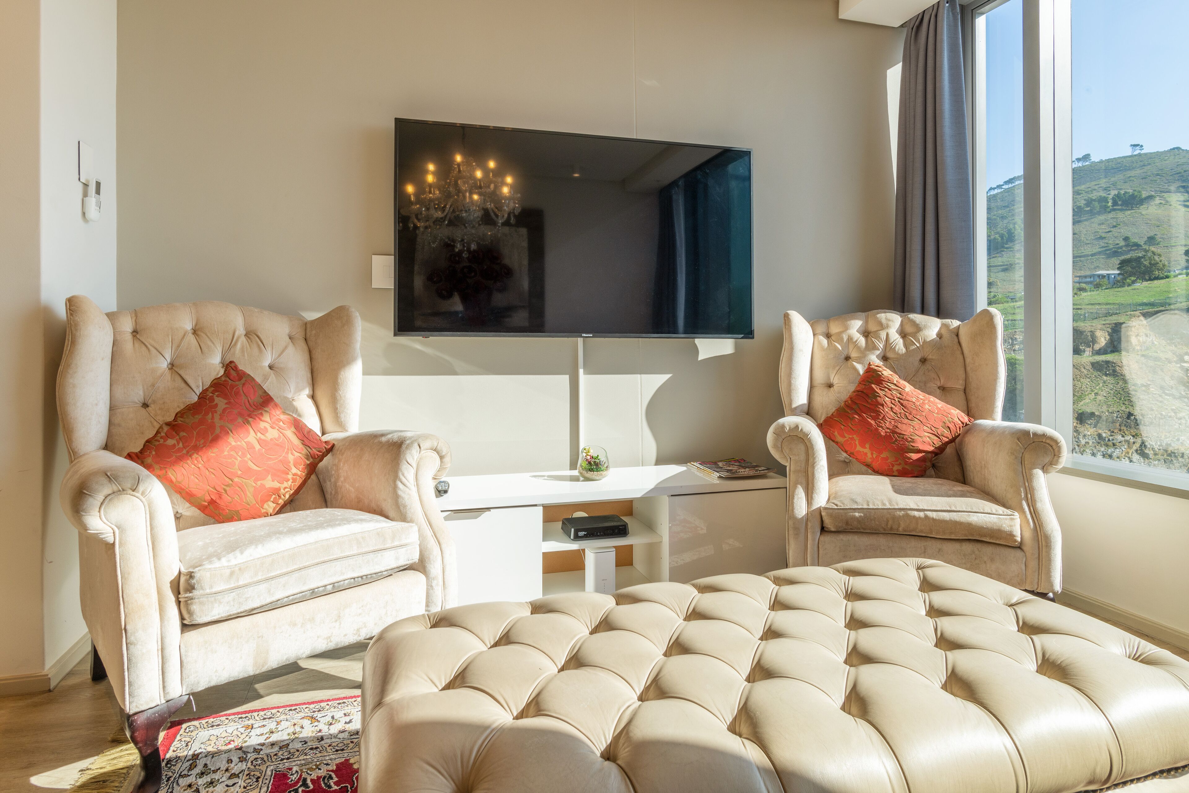 A living room with two armchairs, a large ottoman, a flat-screen TV, and a chandelier.