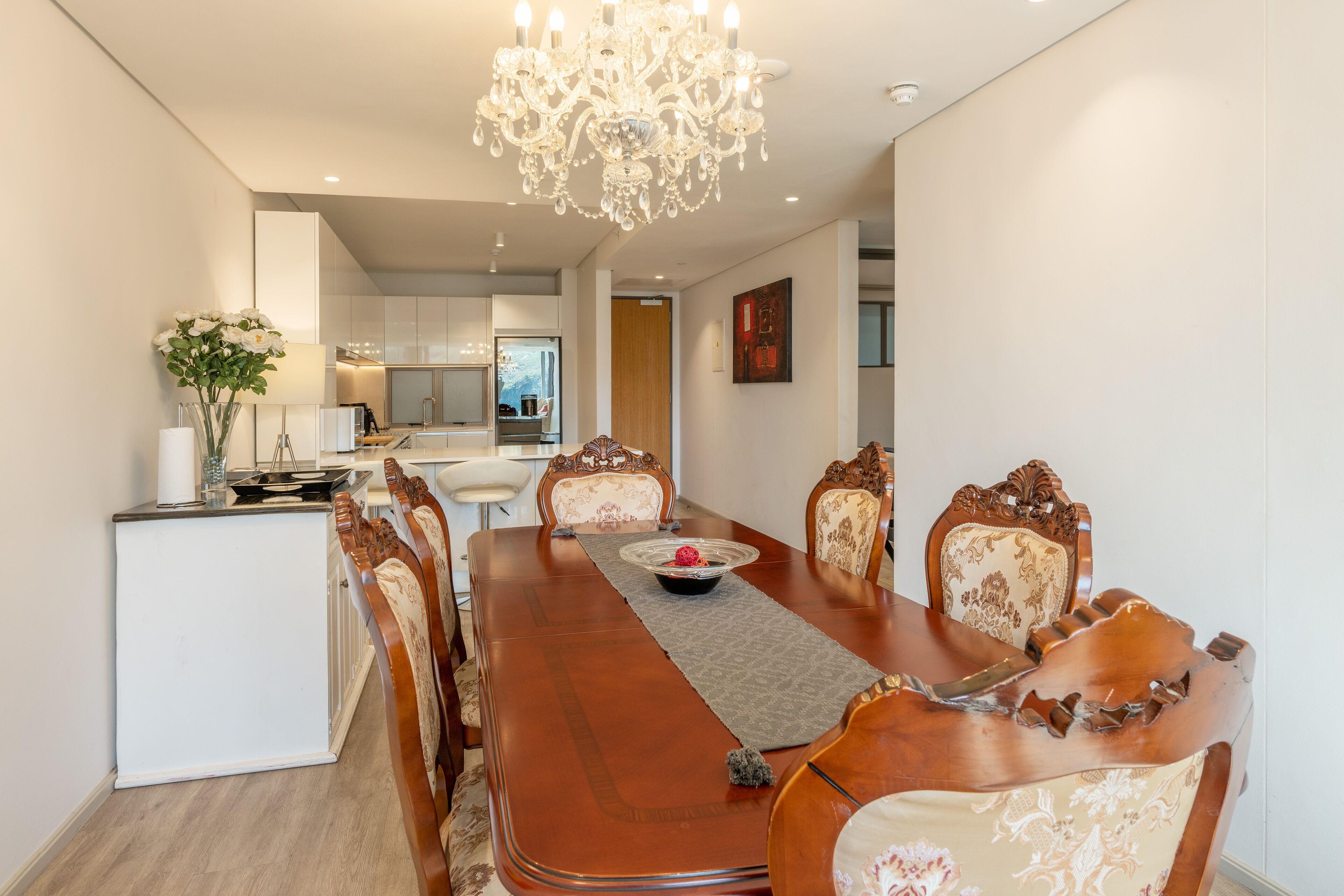 A dining area with a long wooden table, ornate chairs, a chandelier, and a view into the kitchen.