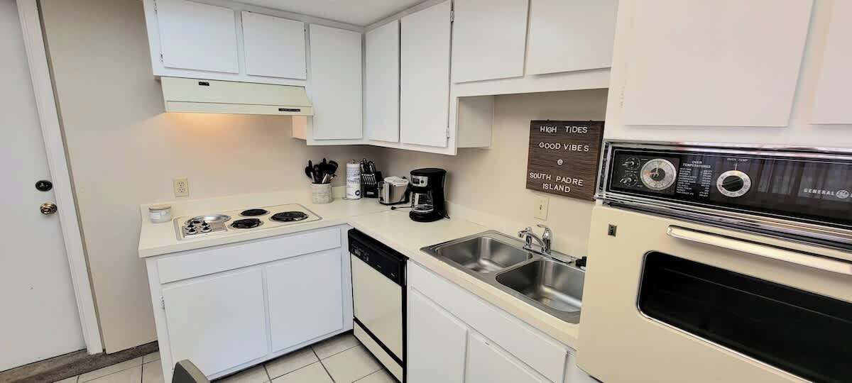 A compact kitchen with white cabinets, a stove, a sink, and a coffee maker.