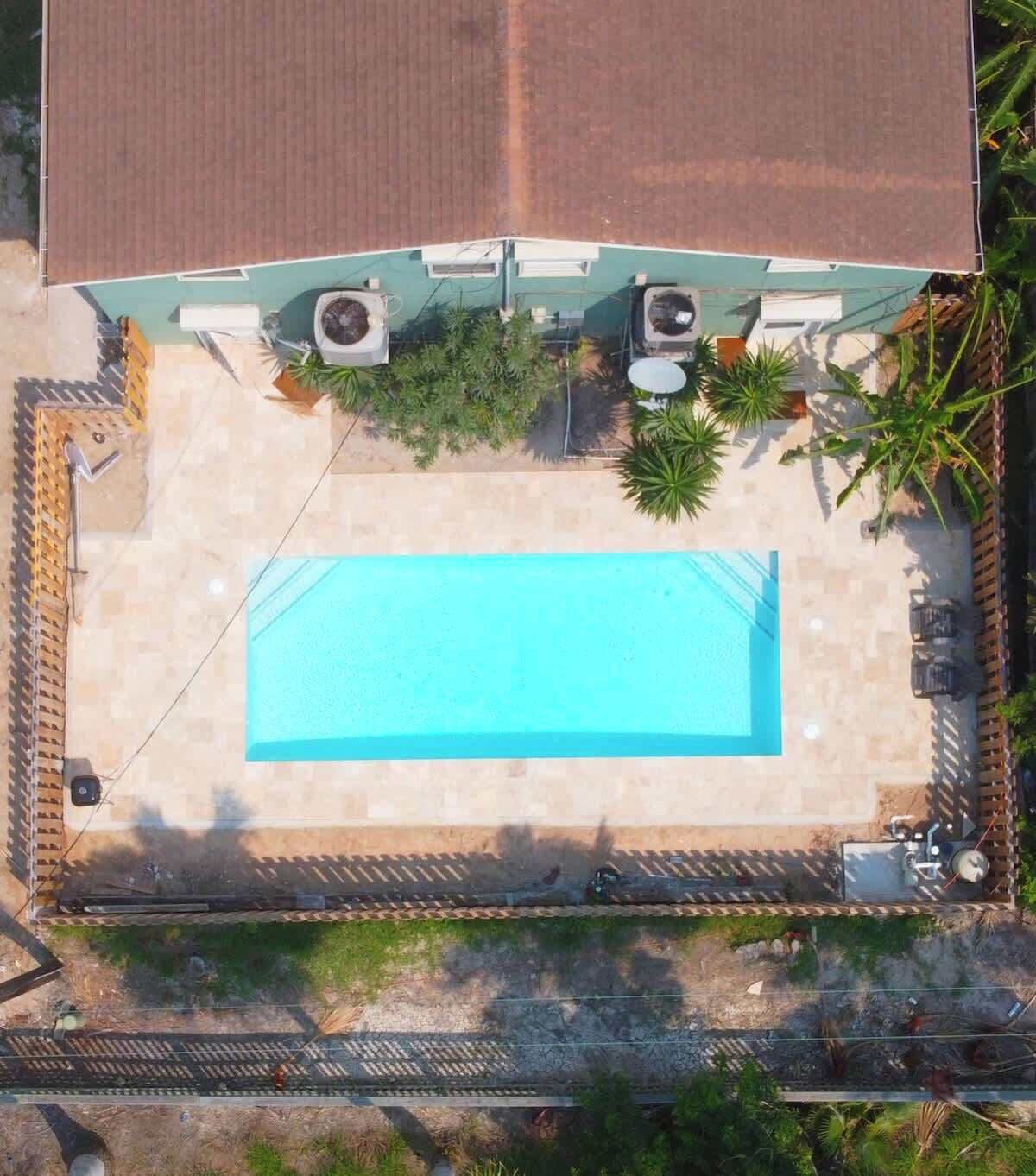 An aerial view of a property with a rectangular swimming pool, a covered patio area, and a fenced garden.