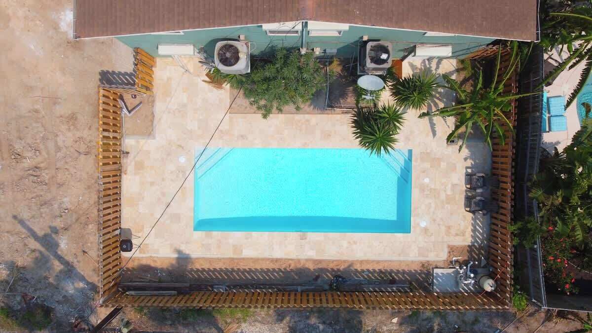 An aerial view of a rectangular swimming pool surrounded by a wooden deck, with two air conditioning units and a small garden area with palm trees.