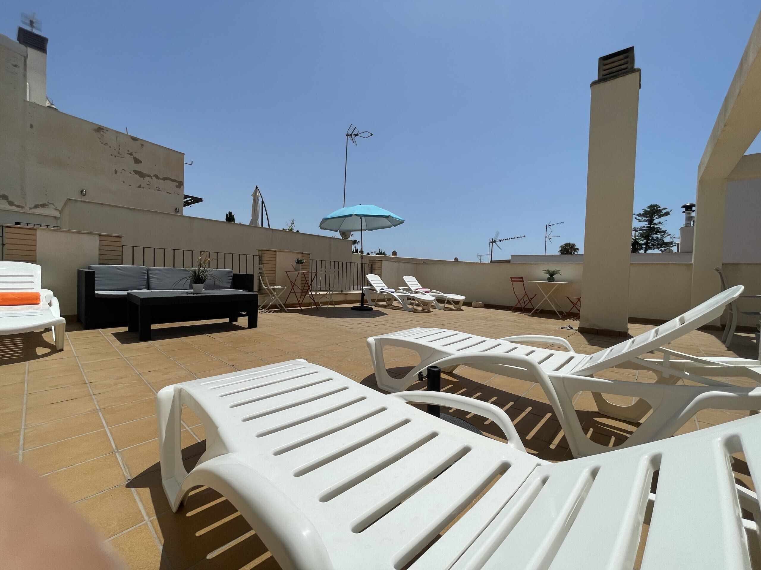 Terraço no telhado com cadeiras de lounge de plástico branco, um sofá preto e um guarda-sol azul.
