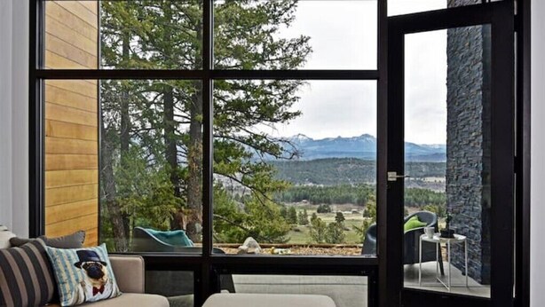 A modern room with a large window offering a mountain view, a sofa with a dog-themed pillow, and a stone accent wall. - Berghaus Pagosa - Experience the Alpenglow (Pagosa Springs)