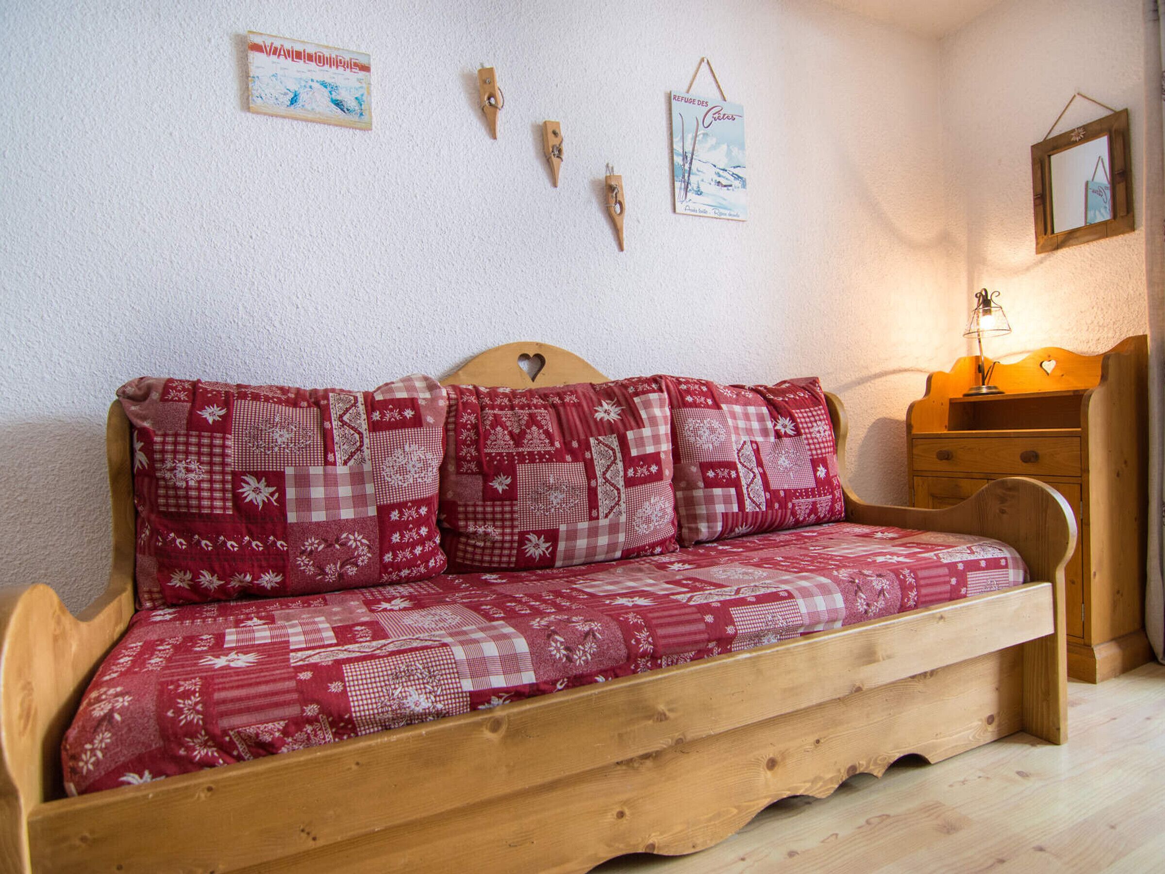 Un canapé-lit en bois recouvert d’un tissu à carreaux rouges et blancs, doté d’une tête de lit en bois et accompagné d’une petite table de chevet en bois avec une lampe.