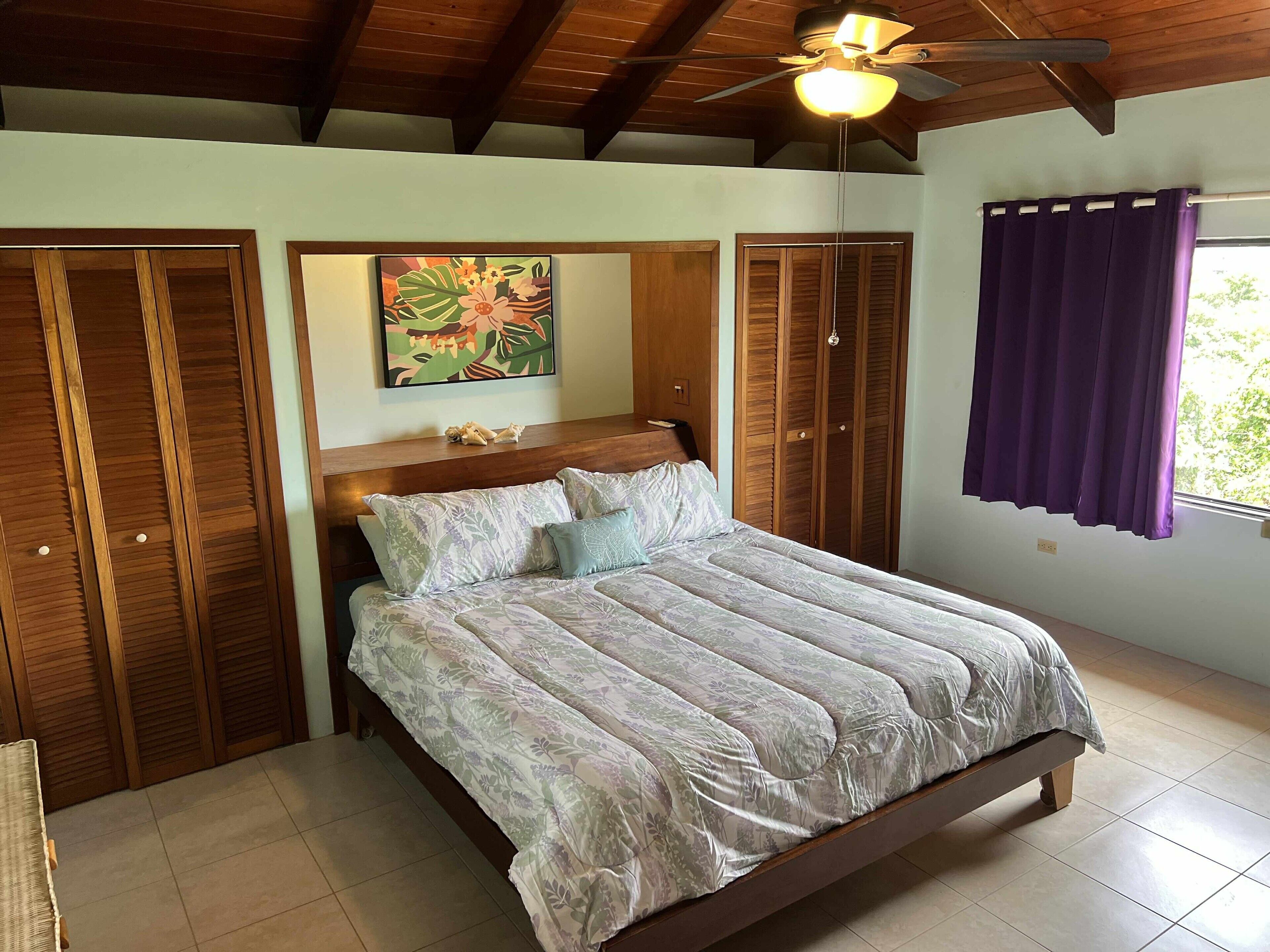 Une chambre à coucher avec un lit, une armoire en bois, un ventilateur de plafond et une fenêtre avec des rideaux.