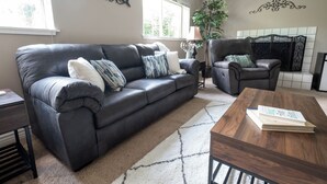 Ein Wohnzimmer mit einem dunkelbraunen Ledersofa, einem Holztisch und einem Sessel. Eine Pflanze, eine Lampe und eine Wanddekoration sind ebenfalls zu sehen.