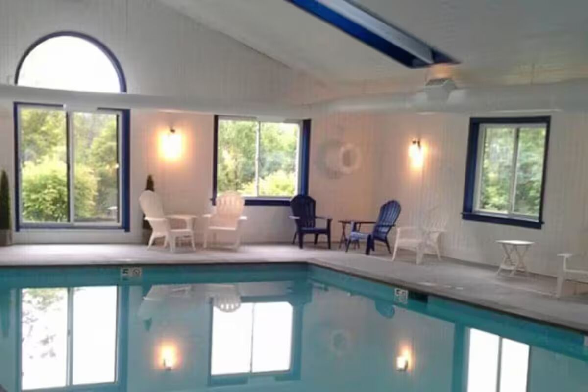 Une piscine intérieure à l’eau d’un bleu cristallin, entourée de murs blancs et de grandes fenêtres donnant sur un paysage verdoyant.