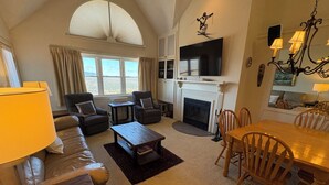 A spacious living room with a fireplace, a large TV, and a dining area with wooden chairs. - Ski In/Ski Out Winterplace 2BR+Loft W/Fireplace, Hot Tub and Pool Access (Ludlow)