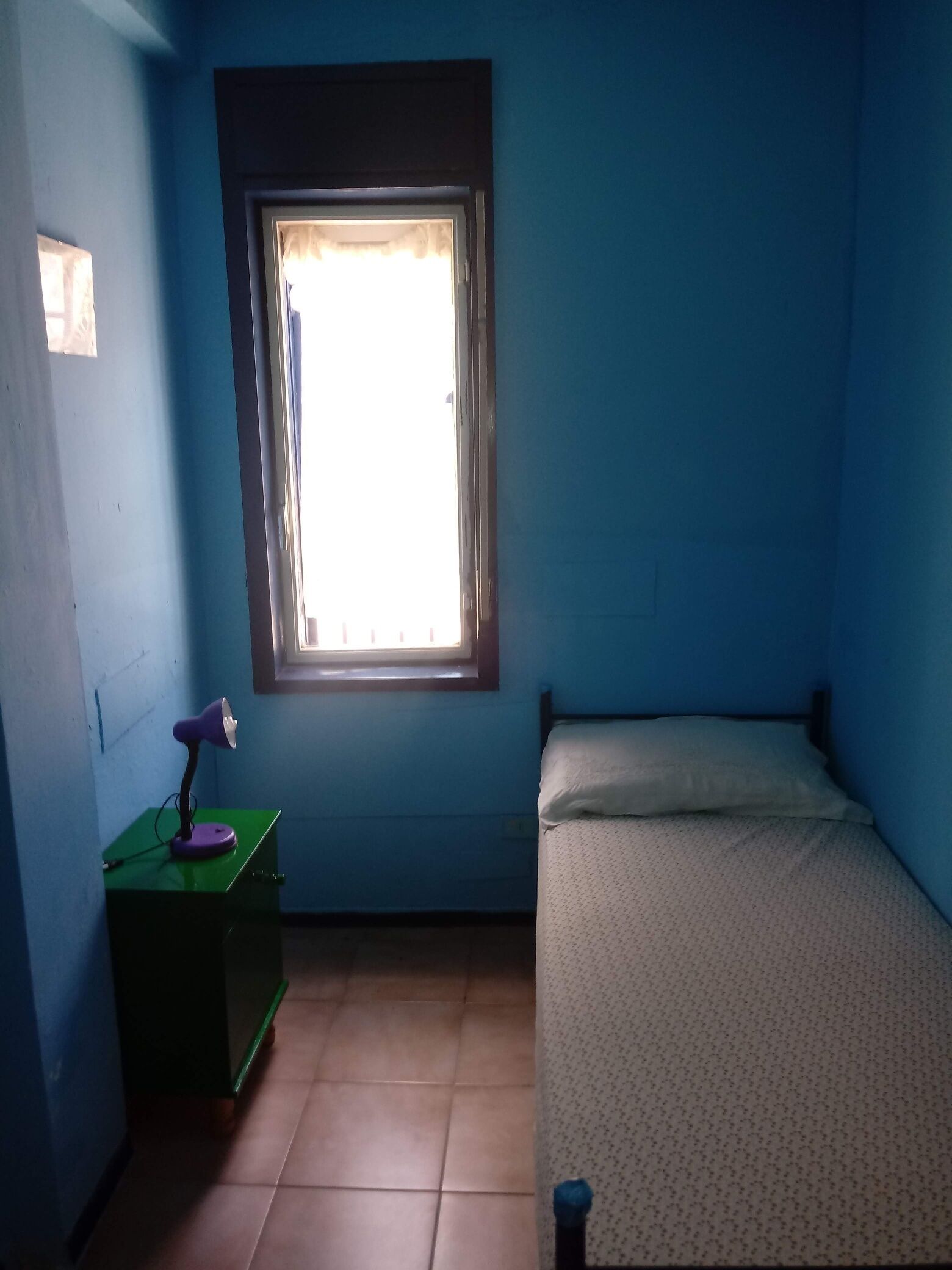 Ein kleines Zimmer mit einem Einzelbett, einem Schreibtisch mit Lampe und einem Fenster mit Jalousien.
