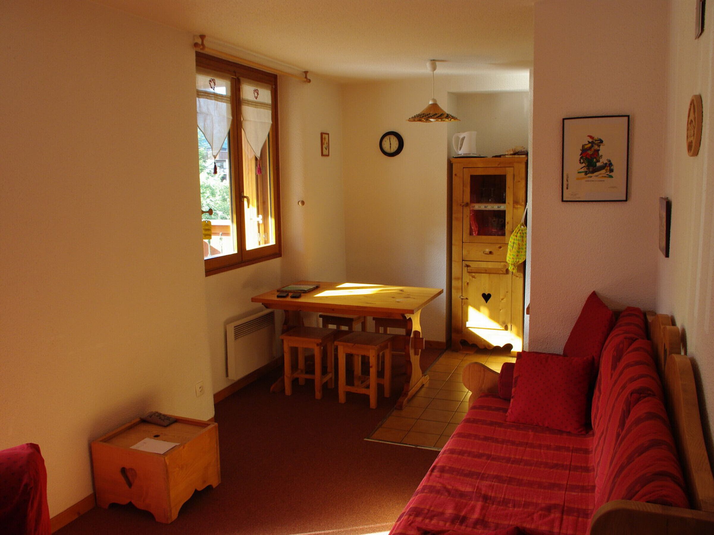 A cozy living room with a red sofa, a wooden dining table with chairs, a kitchen area with a cabinet, and a window with curtains.