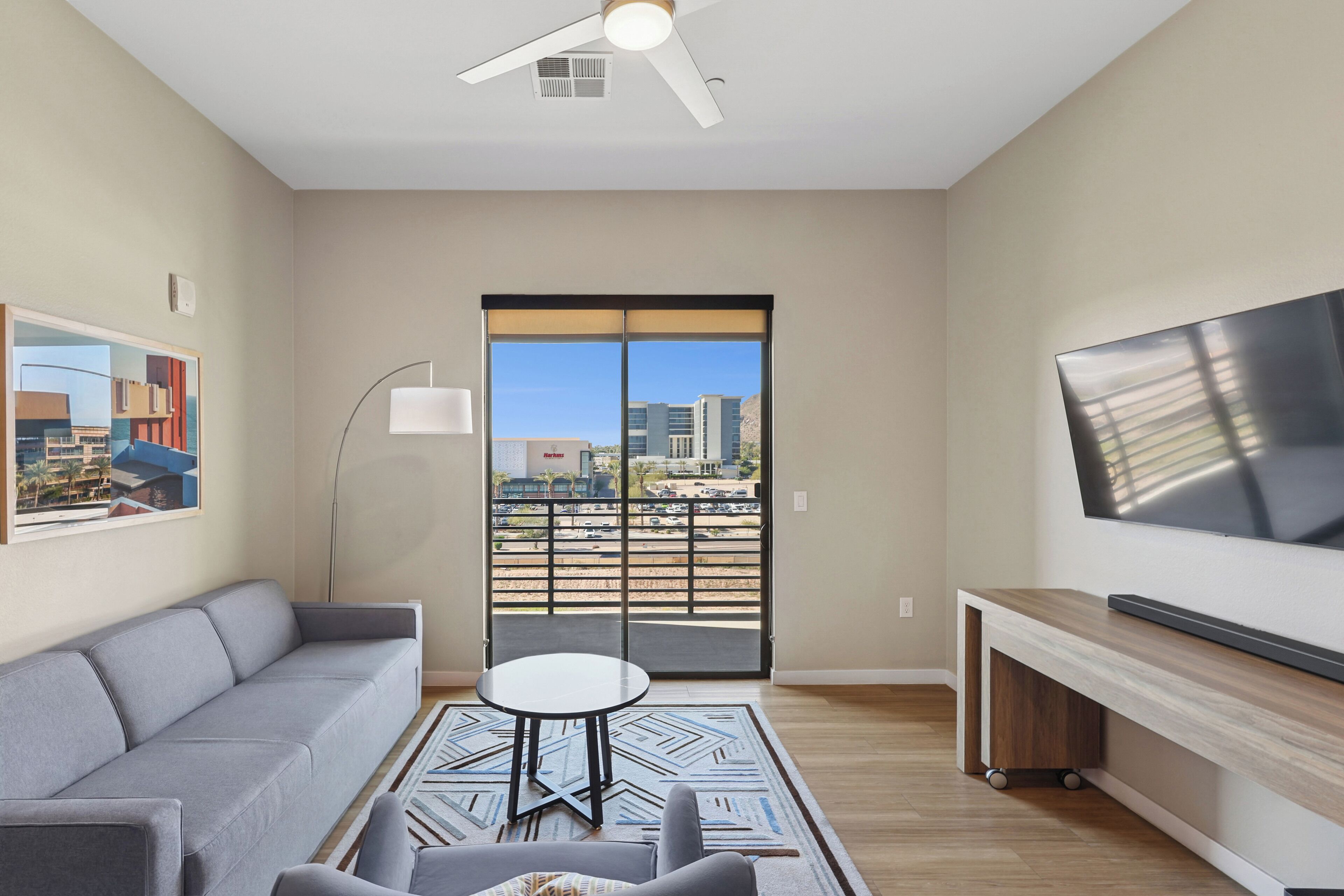 A modern living room with a gray sofa, a round coffee table, and a flat-screen TV mounted on the wall. A large window with a view of buildings and a balcony is visible.